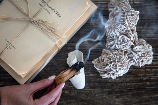 A hand holds a burning Palo Santo stick near a book, sage bundles, and a Desert Rose crystal cluster on a wooden surface.
