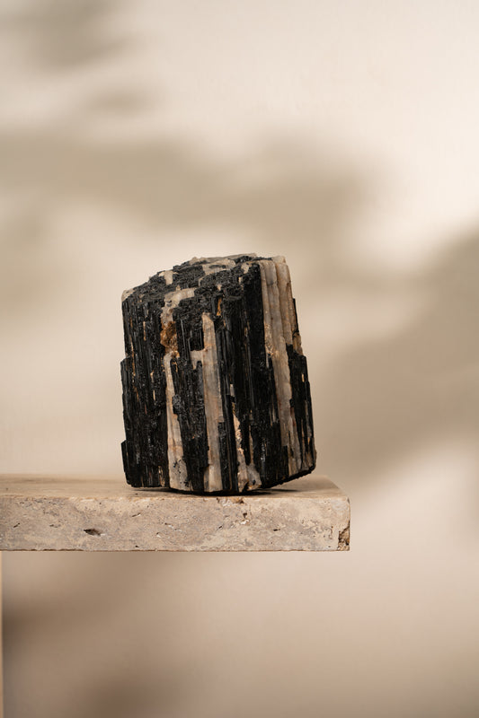 A raw Black Tourmaline crystal cluster with distinct black striated formations, contrasting with light-colored mineral inclusions, resting on a neutral stone base.
