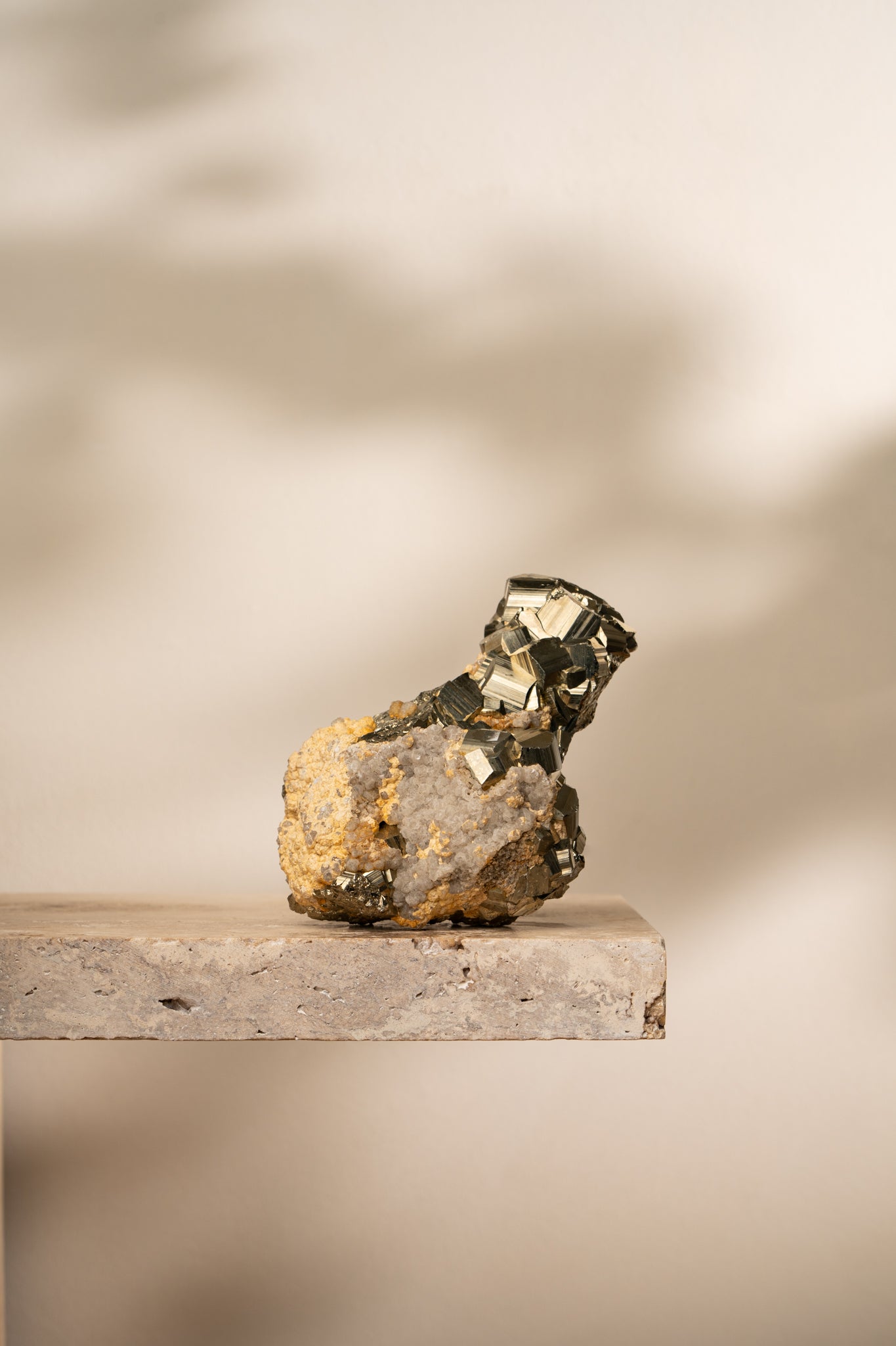 A raw Pyrite formation with bright metallic gold cubic structures emerging from a natural earthy-toned mineral matrix, resting on a neutral stone surface.