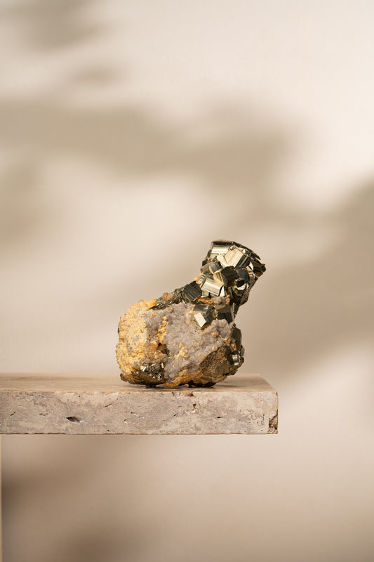 A raw Pyrite formation with bright metallic gold cubic structures emerging from a natural earthy-toned mineral matrix, resting on a neutral stone surface.
