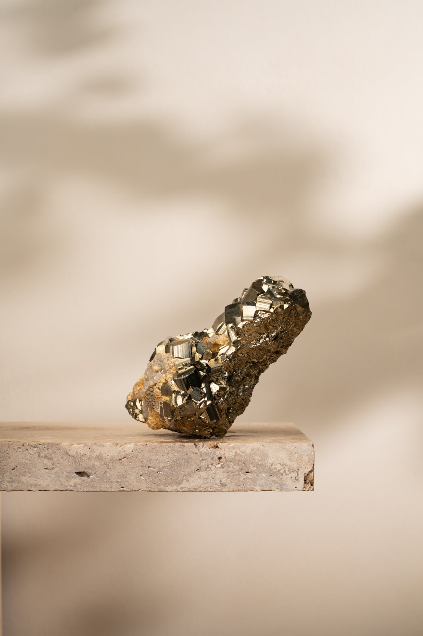 A raw Pyrite formation with bright metallic gold cubic structures emerging from a natural earthy-toned mineral matrix, resting on a neutral stone surface.