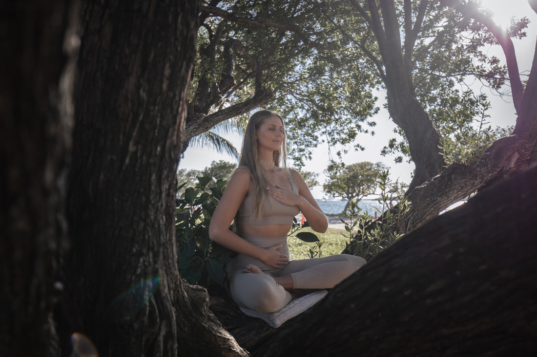 Woman meditating outdoors, seated between tree roots with a serene seascape in the background.