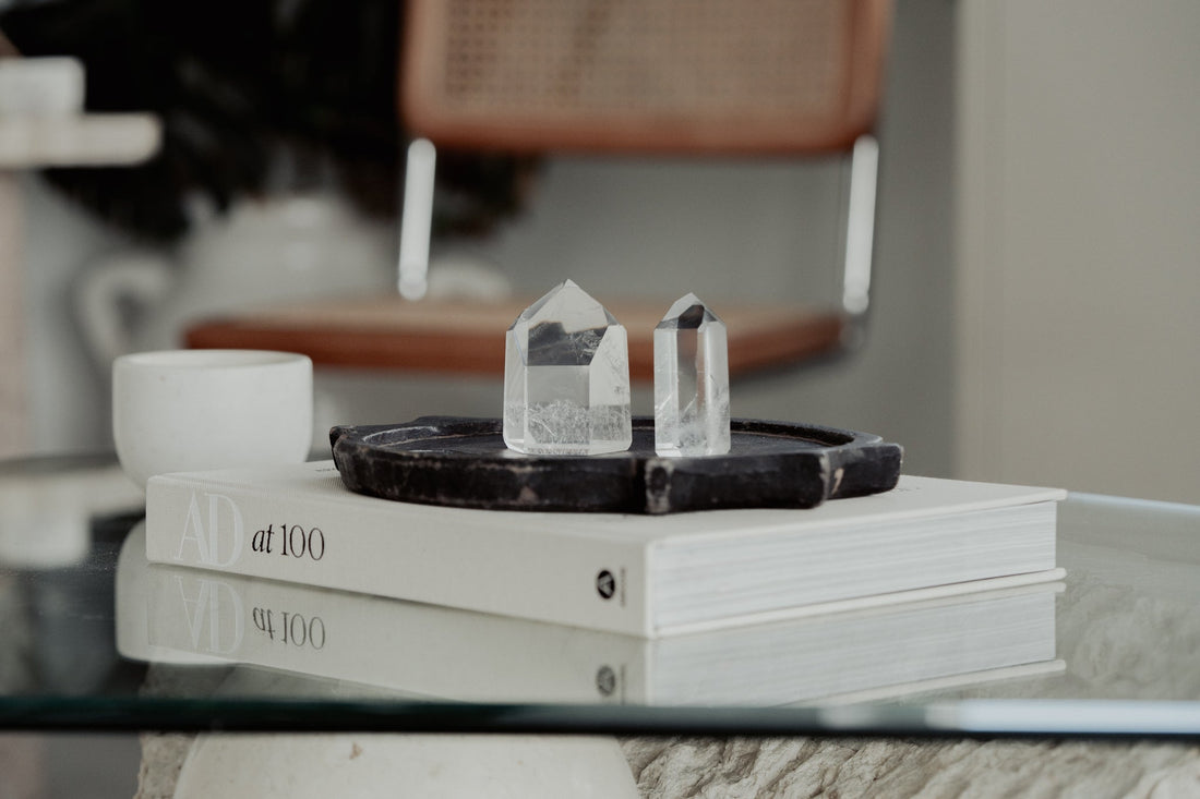 Two clear quartz crystals on a black tray, atop design books on a glass table in a serene setting.
