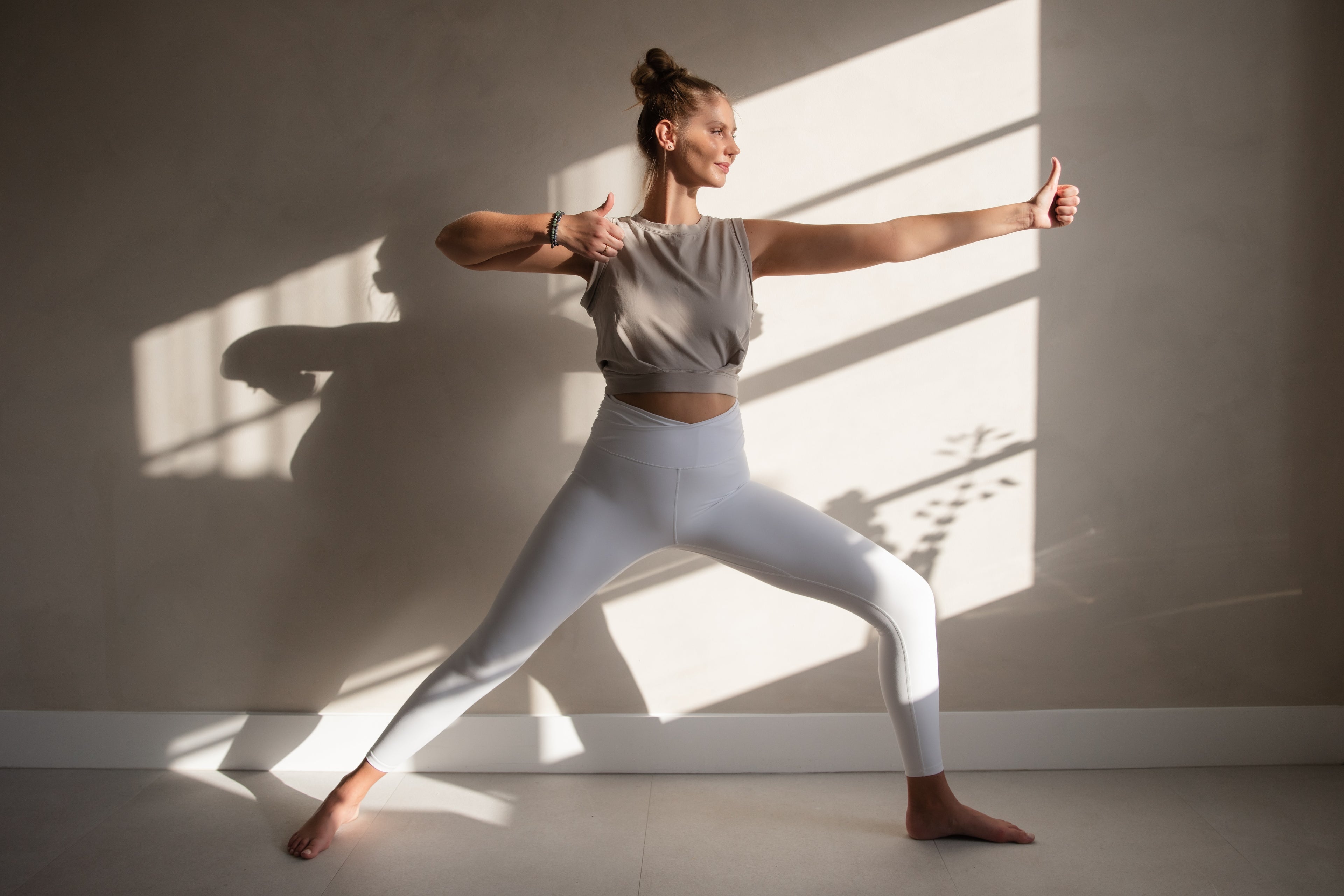 Woman in gray athletic wear standing in a yoga pose with sunlight casting shadows on a wall.