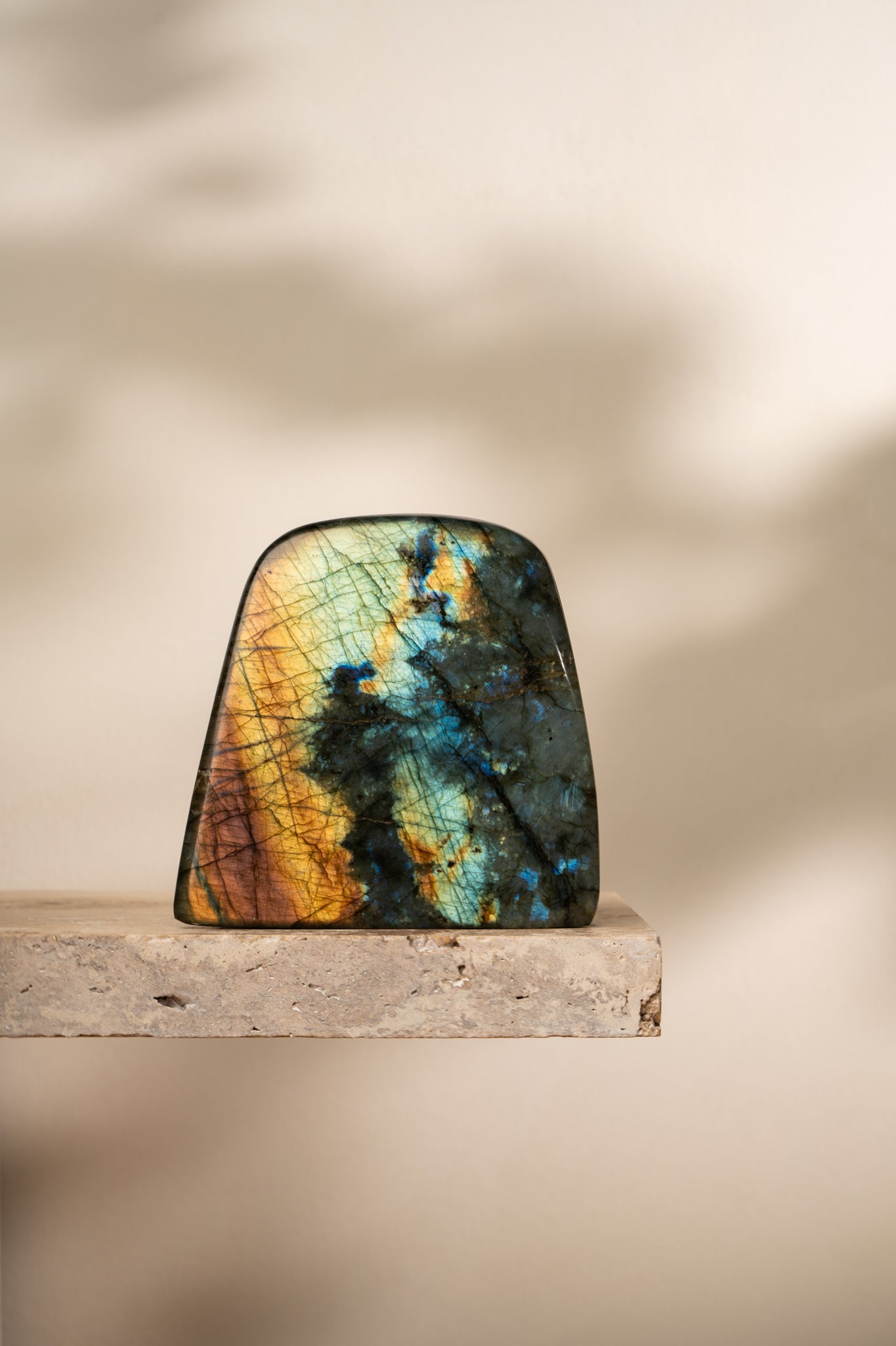 A standing polished Labradorite crystal with flashes of blue, gold, and green against a dark matrix, positioned on a stone slab with soft shadows.