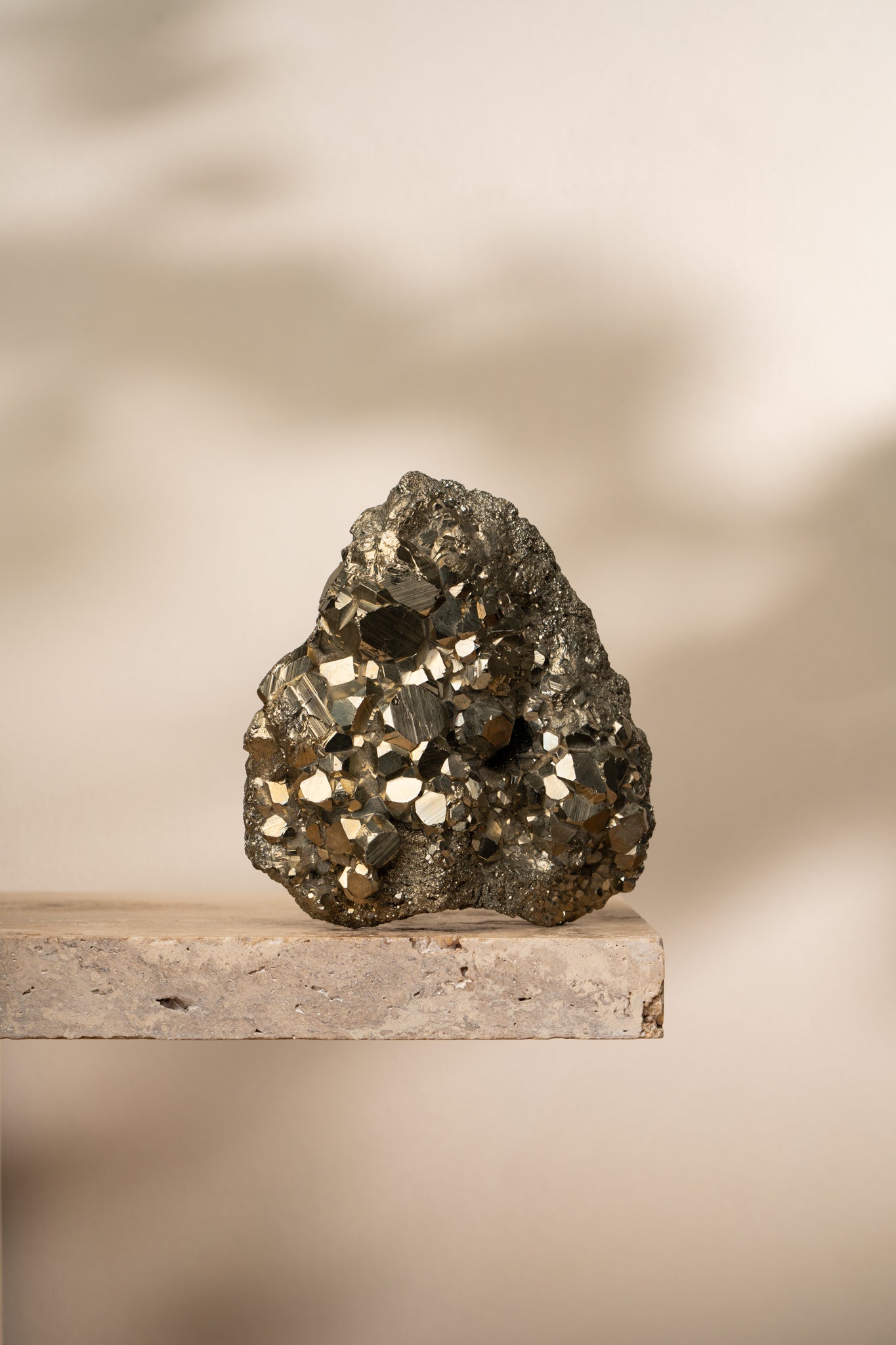 A large natural Pyrite cluster with a metallic gold luster, featuring sharp geometric formations. The raw crystal sits on a neutral stone shelf, reflecting light beautifully.