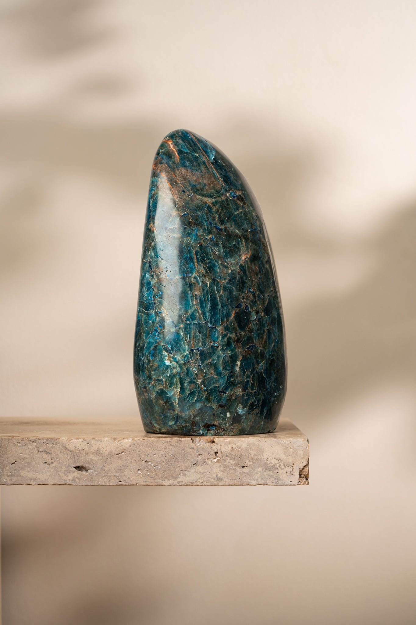 A polished Blue Apatite freeform crystal with deep blue and green hues, standing upright on a neutral stone surface. Its smooth surface reflects light beautifully.